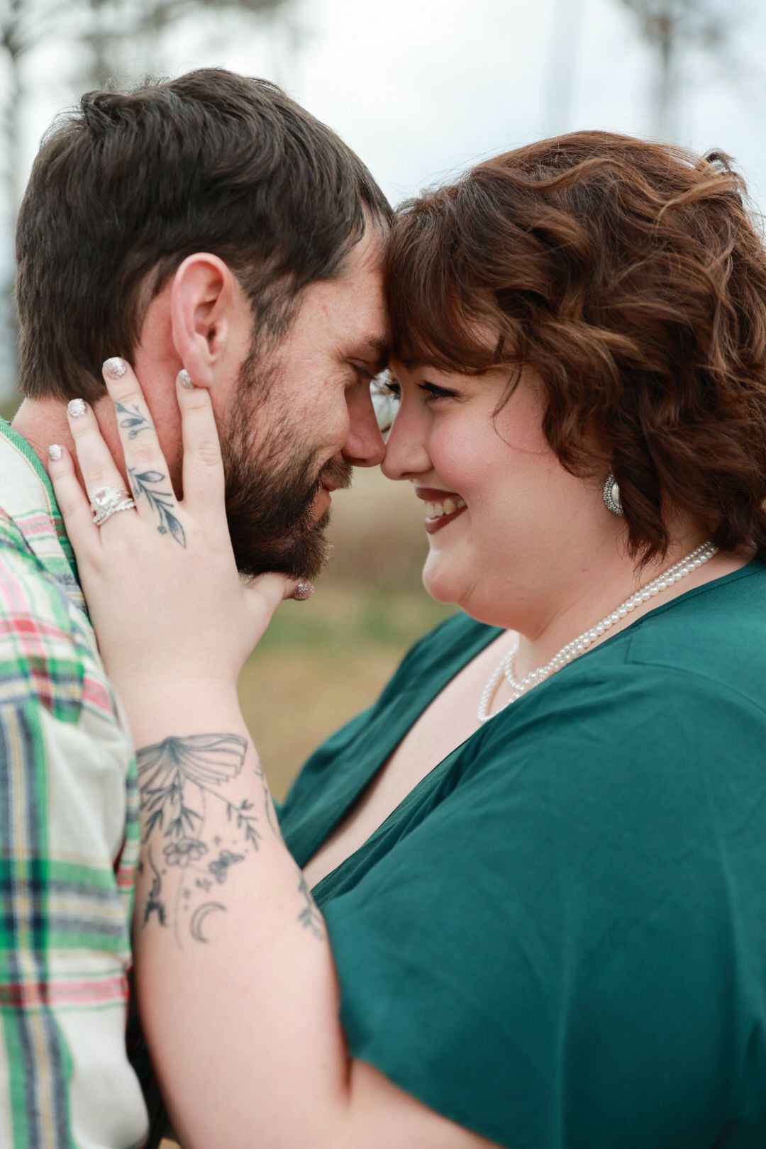 Bearded man with tattooed partner in a teal dress touches foreheads and smiles during a romantic outdoor engagement session.