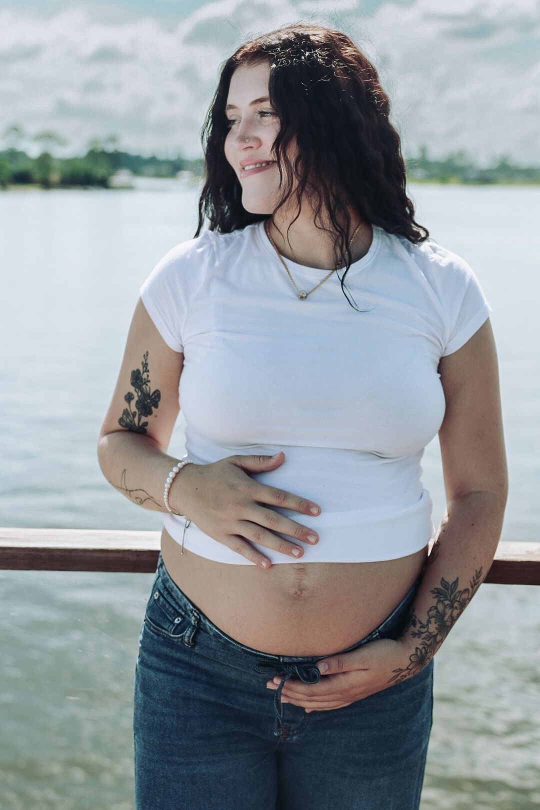 Pregnant woman with dark wavy hair and tattoos wears a white tee and jeans, holding her baby bump beside a lake under cloudy skies.