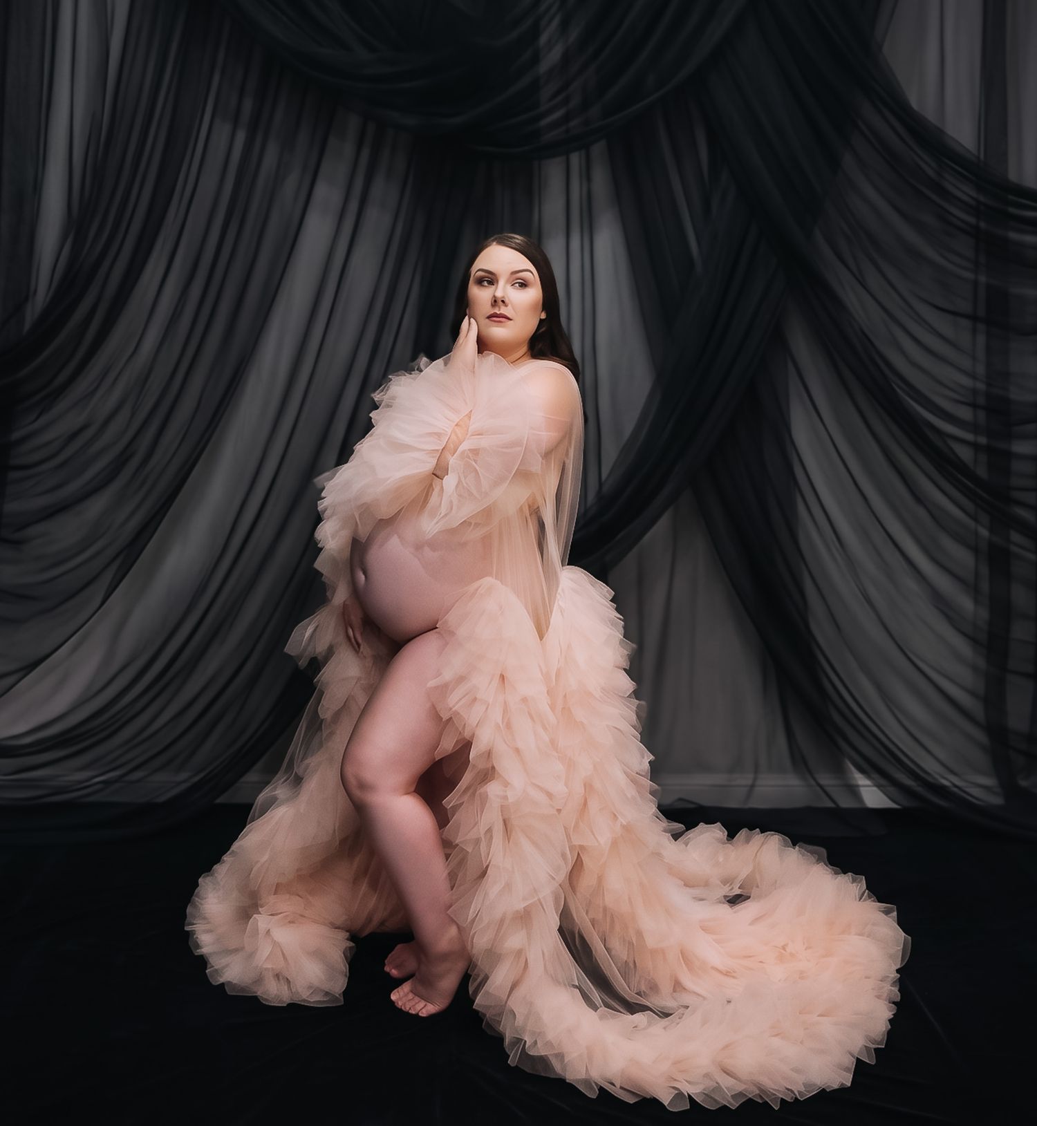 Pregnant woman poses in a blush ruffled tulle gown cradling her belly in front of flowing black sheer drapes in a studio.