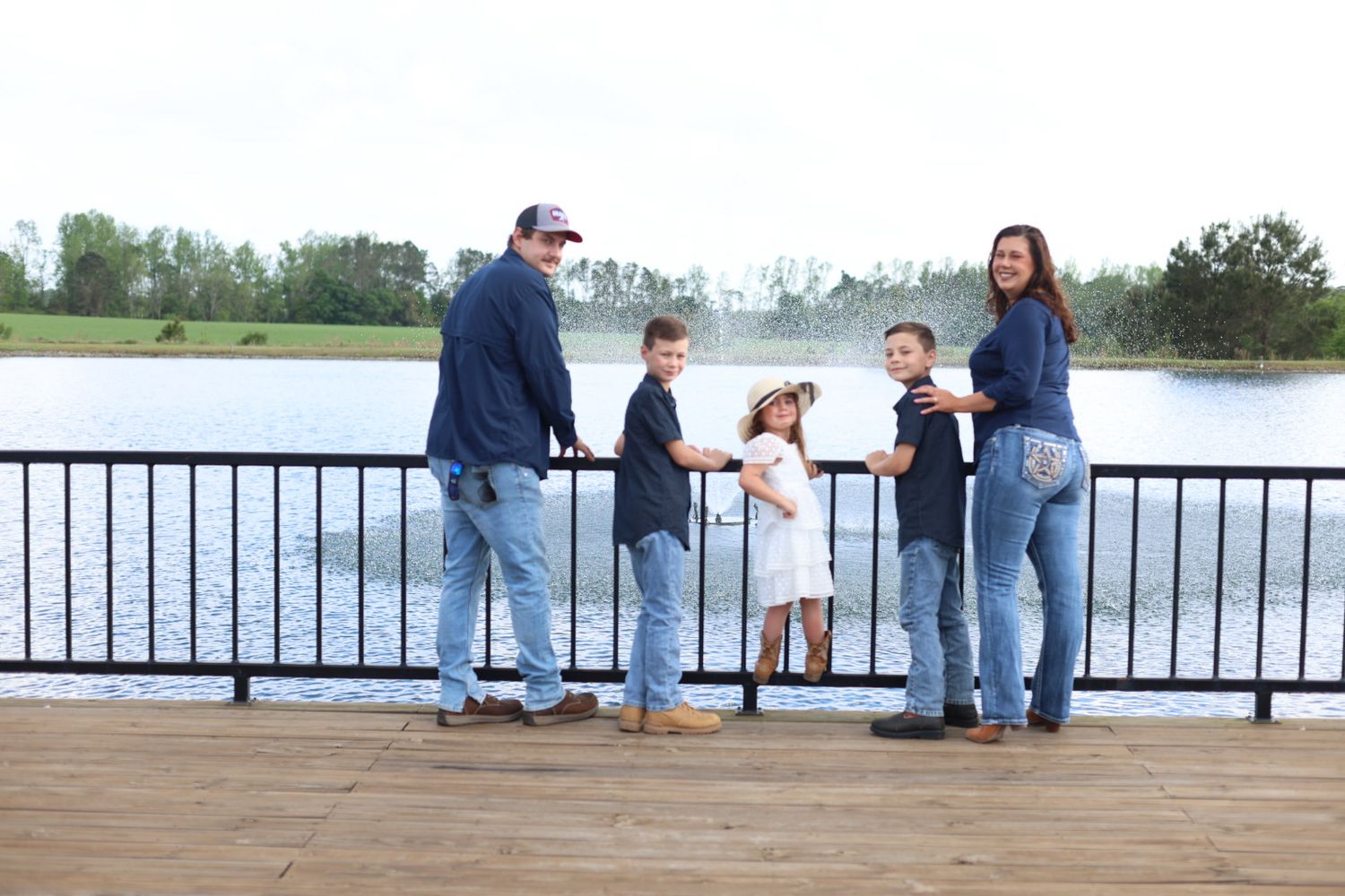 Family of five standing at a black railing on a wooden dock, facing a lake with a fountain on a sunny day.