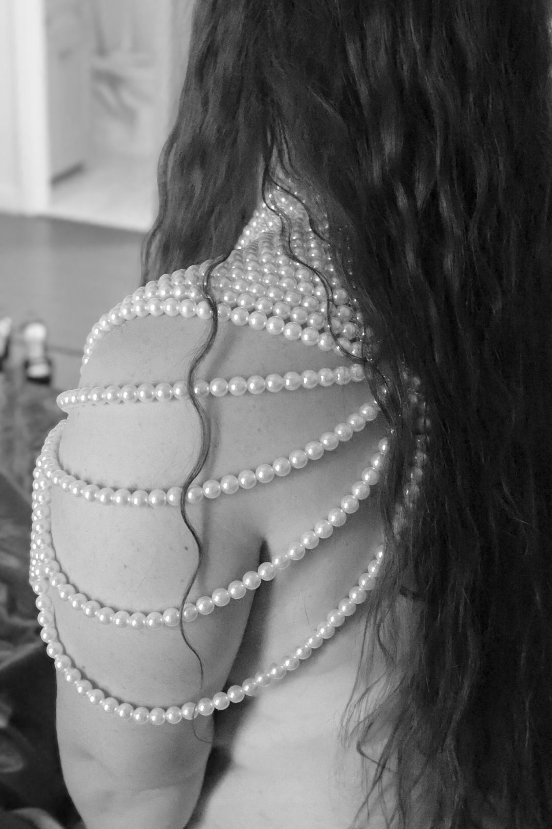 Black and white close-up of a woman's shoulder draped in layered pearl strands with long wavy dark hair falling down her back.