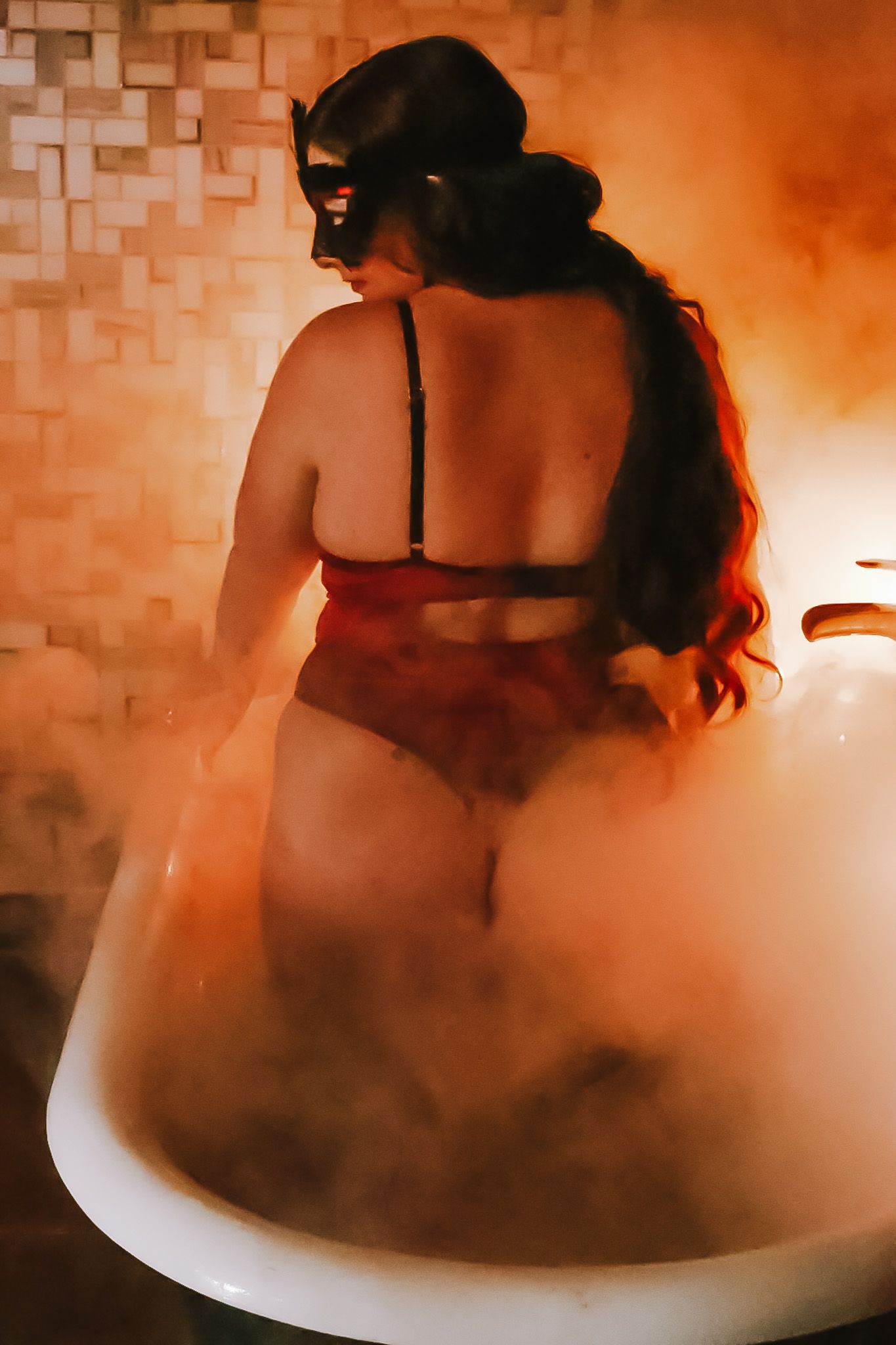 Woman in a mask and red lingerie sits in a vintage clawfoot tub, surrounded by orange glowing steam and tiled walls.