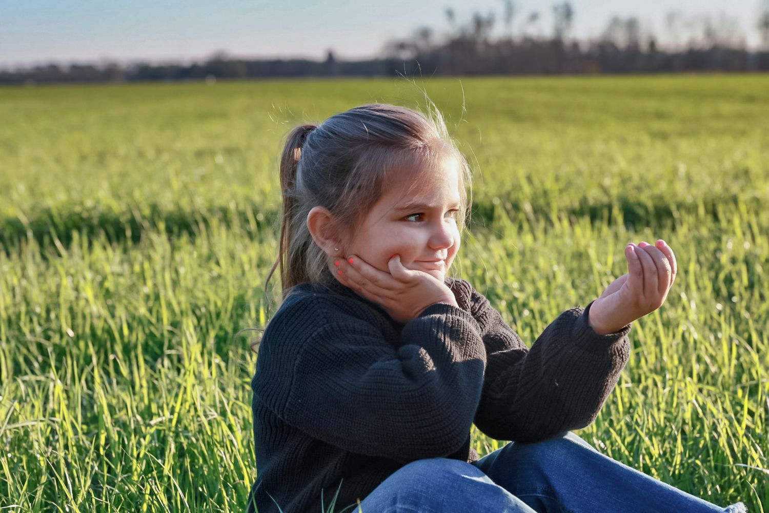 Young girl with a ponytail sits in a green grass field, resting her cheek on her hand in warm evening light.