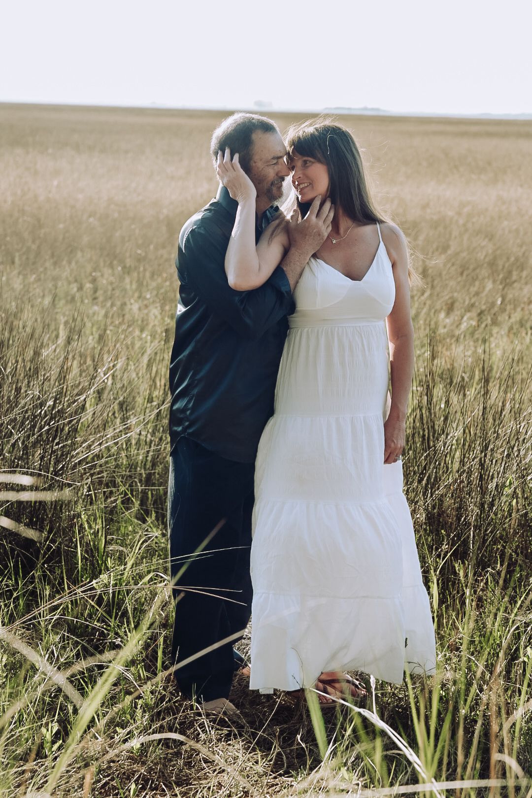 Couple in white dress and dark shirt embrace tenderly while standing in a tall golden marsh grass field.