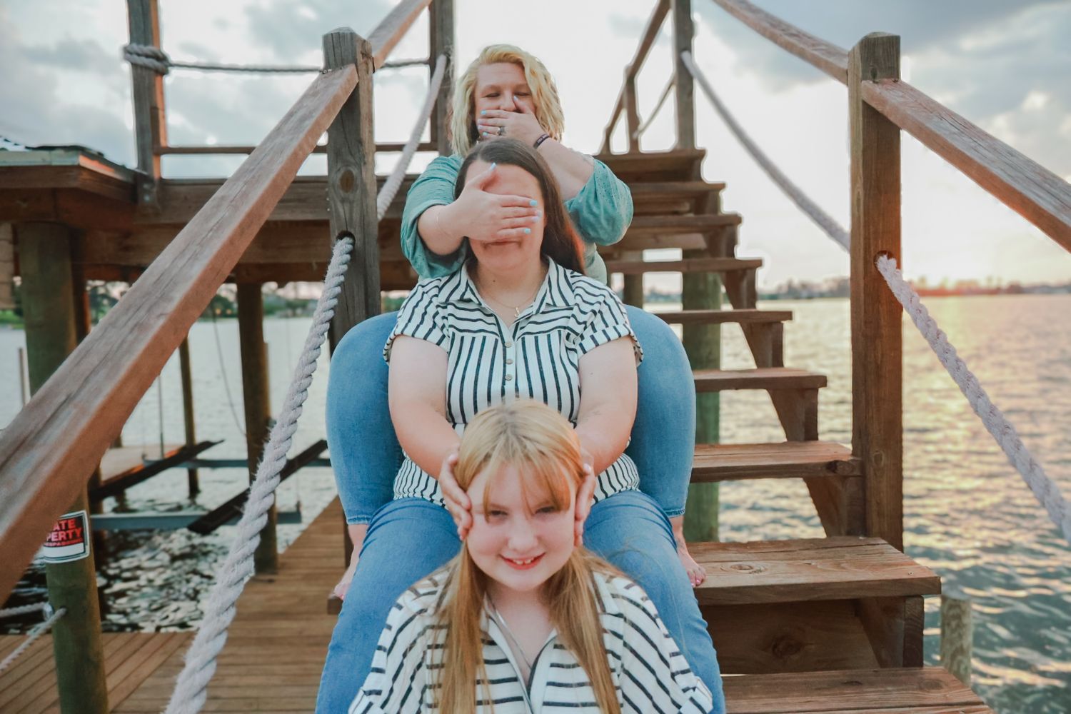Two women and a young girl pose on wooden dock steps by the water at sunset, one woman playfully covering another's eyes.