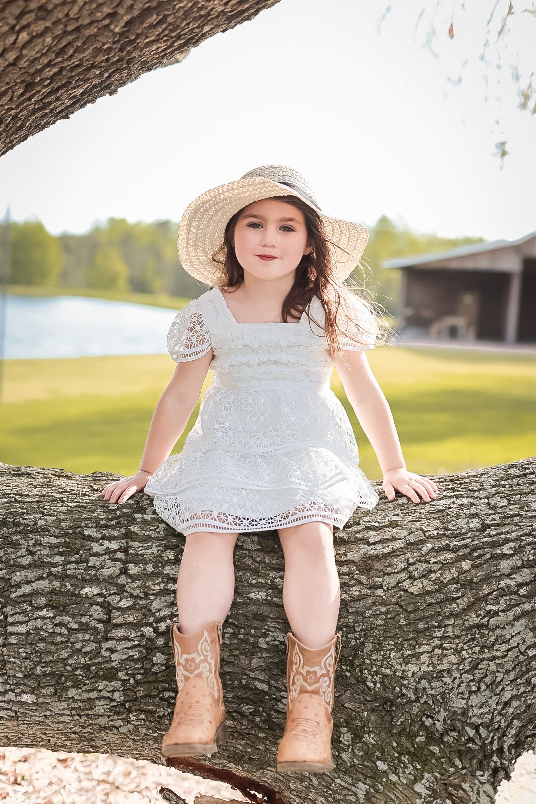 Young girl in a white lace dress, straw hat, and tan cowboy boots sits on a large oak tree branch above a grassy field.