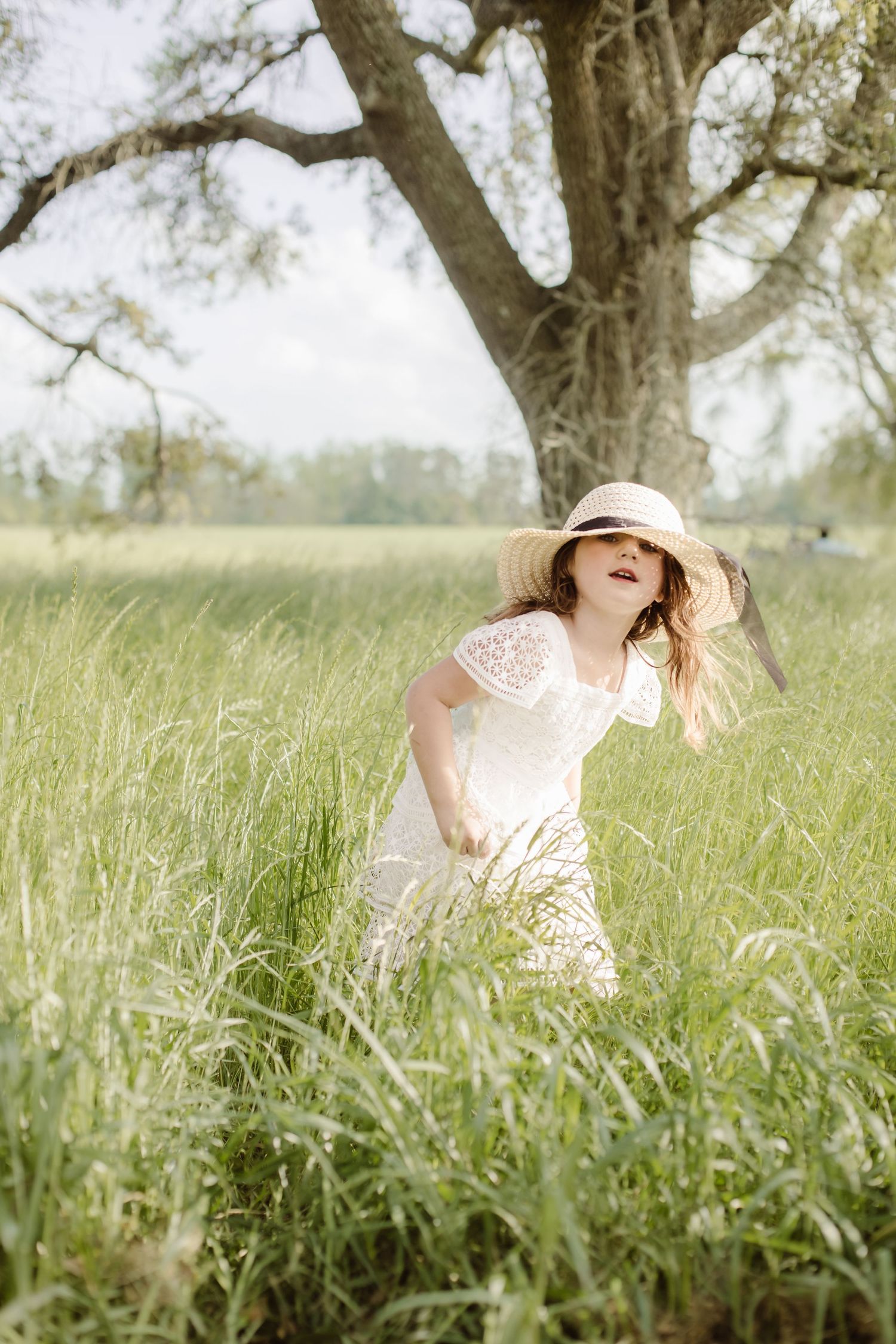 Playful young girl in a white lace dress and straw hat running through tall wild grass beneath a large oak tree.