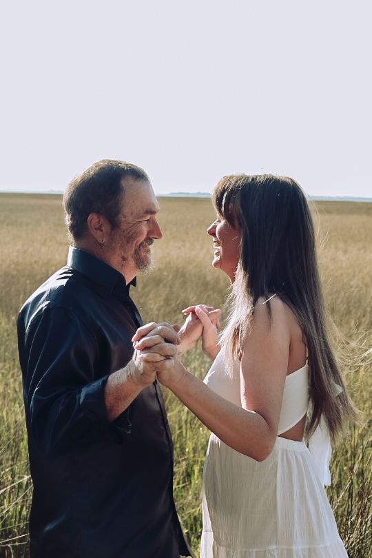 Smiling couple hold hands and gaze at each other in a open field of golden grass under a bright sky.
