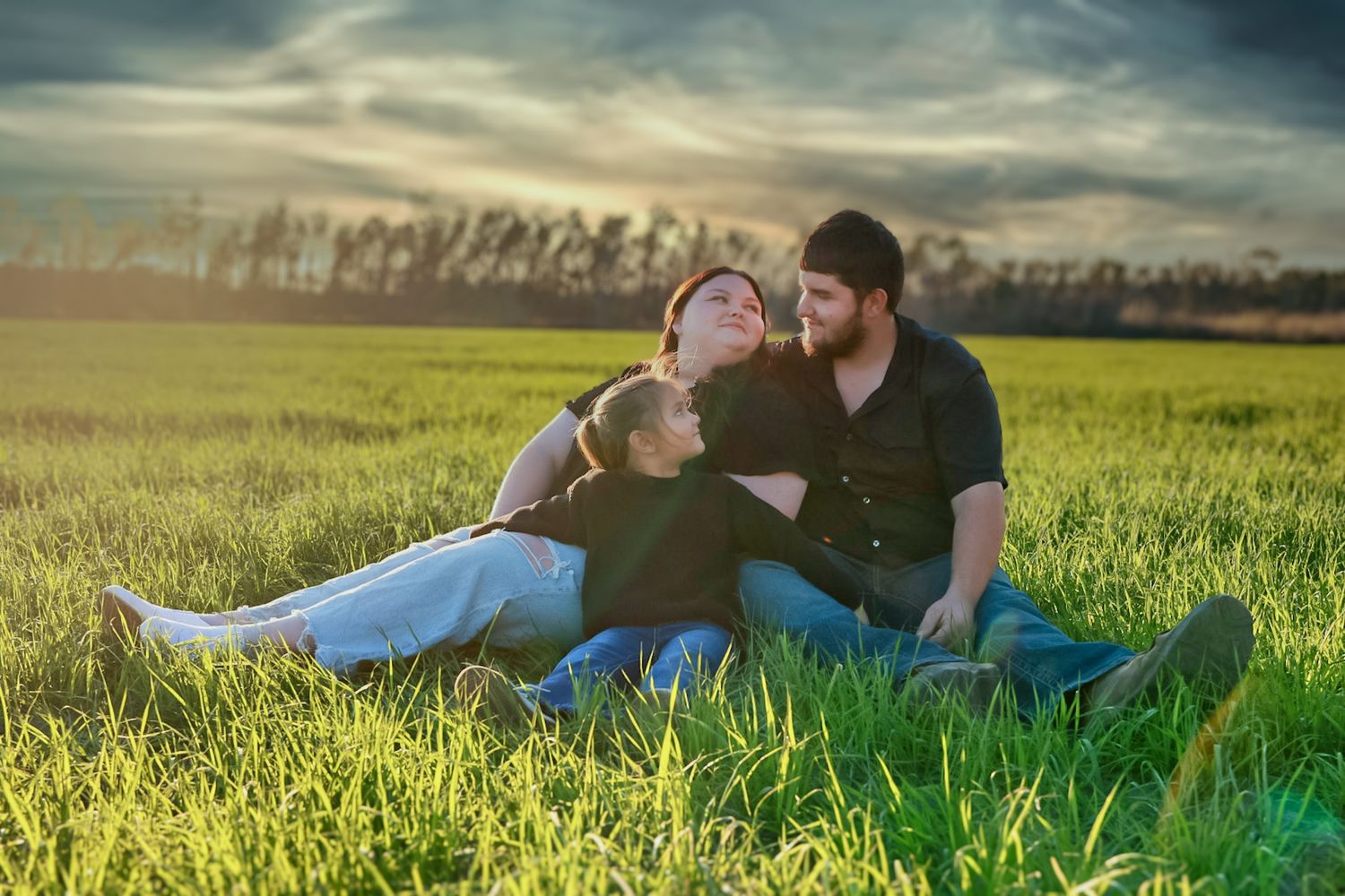 Couple and their young daughter relax together in a lush green field under a dramatic sunset sky.