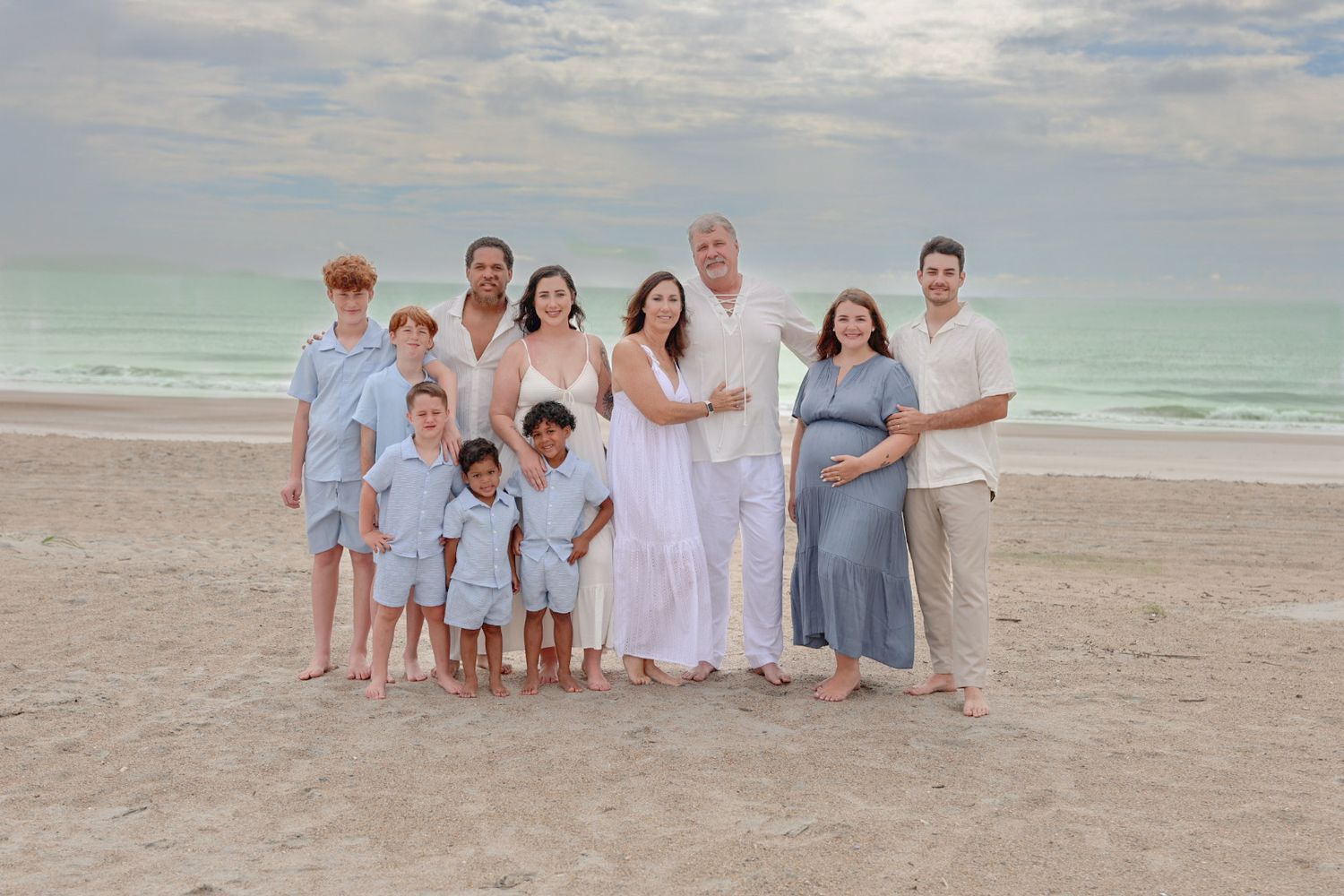 Multi-generational family in matching blue and white attire standing on the beach at sunset with calm ocean waves behind.
