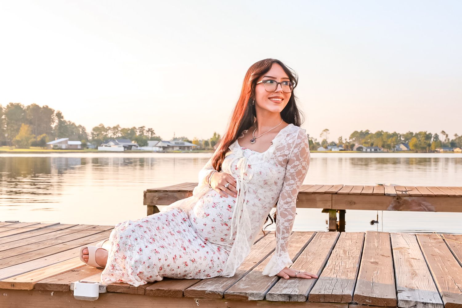 Young woman with long hair and glasses sits on a wooden lakeside dock at sunset wearing a floral white lace dress.
