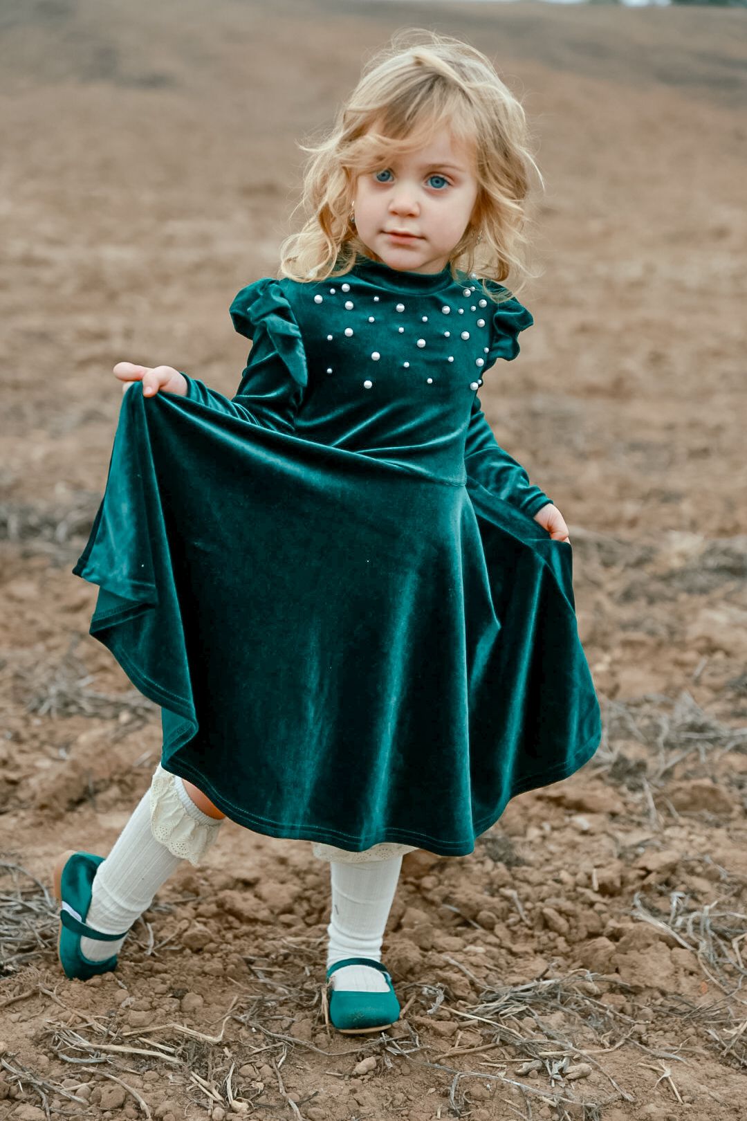 A little girl twirls the skirt of her green velvet dress with pearl details while standing on bare dirt in matching green shoes.
