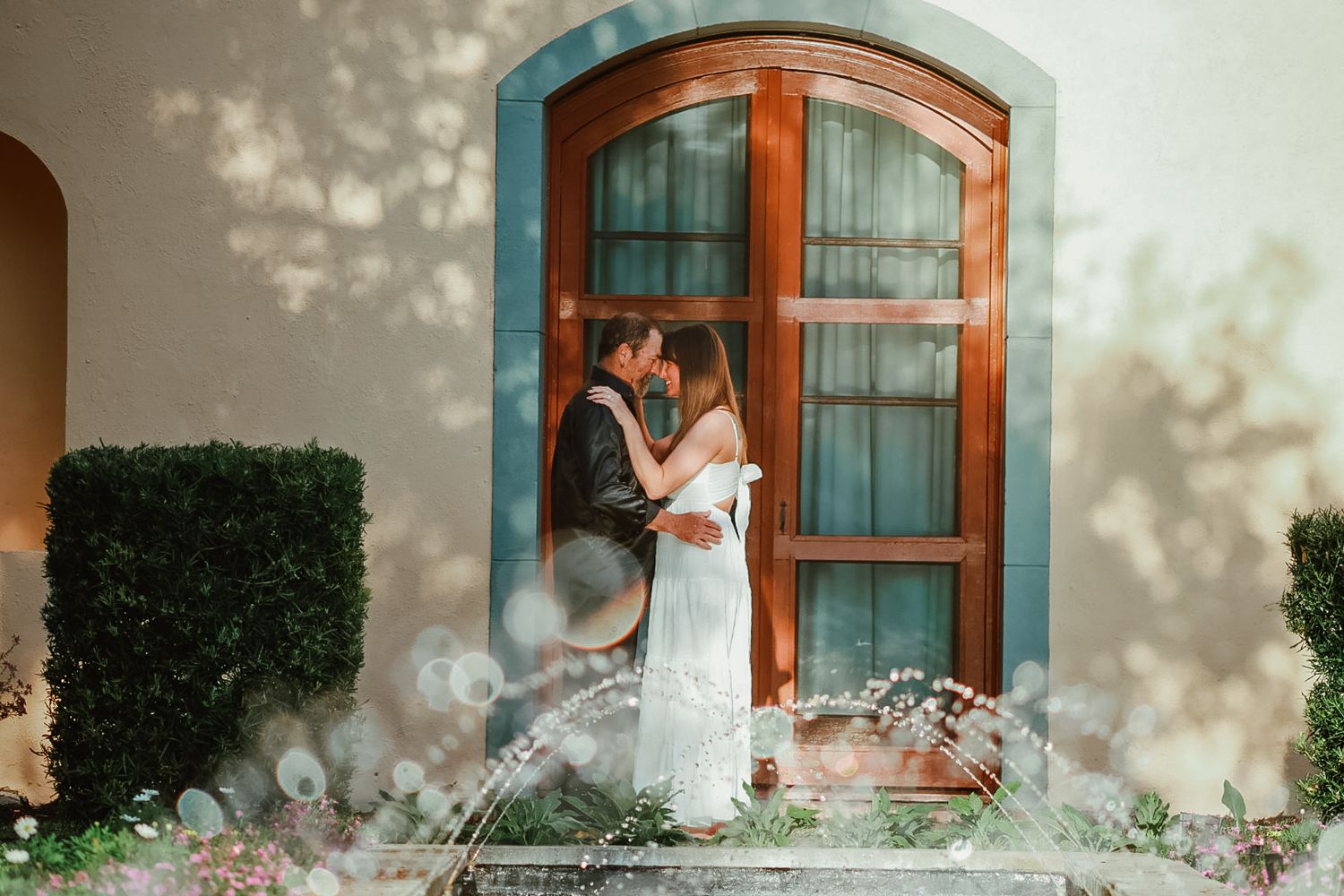 Couple shares an intimate moment in front of an arched wooden window with soft sunlight, shadows, and bubbles in the air.