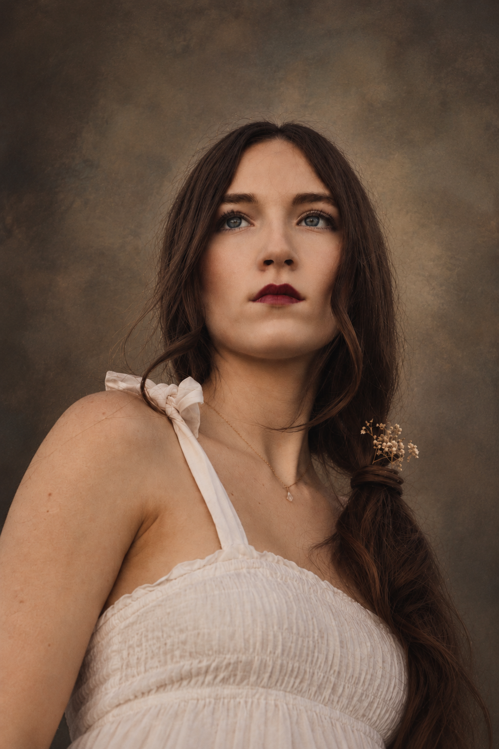 Moody studio-style portrait of a woman with a loose side braid, deep red lipstick and white dress against a textured brown backdrop.