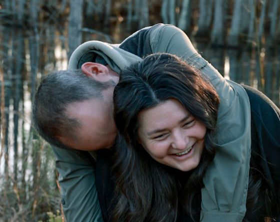 Smiling woman with long dark hair laughs as a man in an olive jacket leans playfully against her in a sunlit woods.