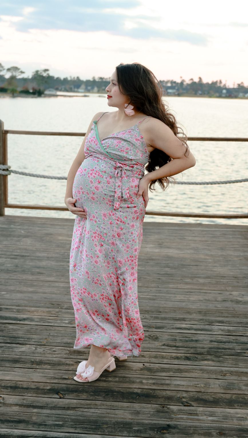 Pregnant woman in a long pink floral dress poses with a hand on her hip on a wooden lakeside dock at sunset.