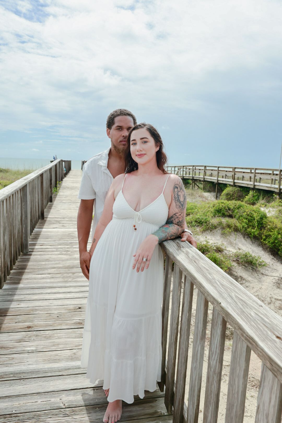 A woman in a white sundress with an arm tattoo stands in front of a man on a wooden beach boardwalk near dunes.