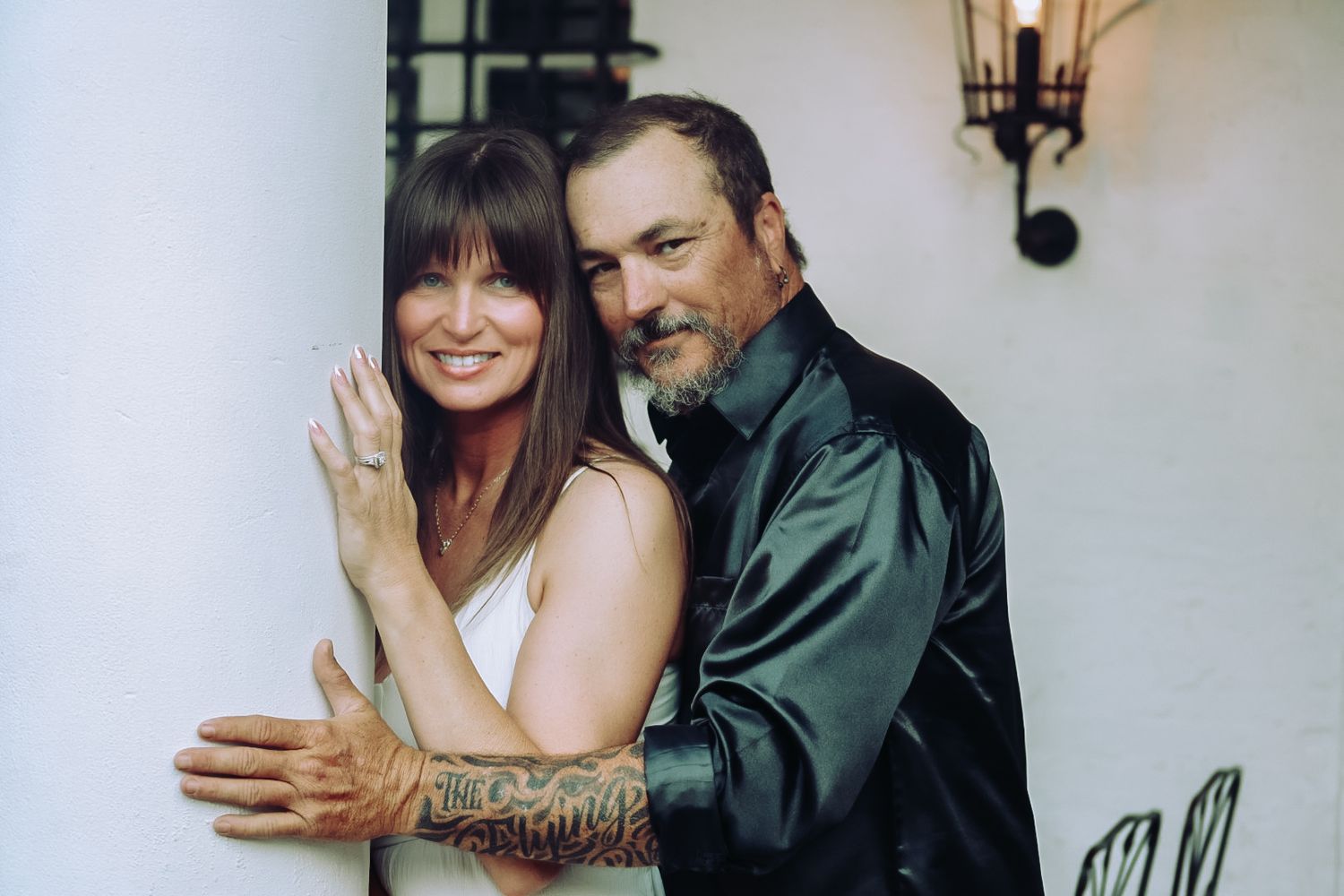 A man with a tattooed arm in a satin black shirt hugs a smiling woman with bangs beside a white wall and wall lantern.