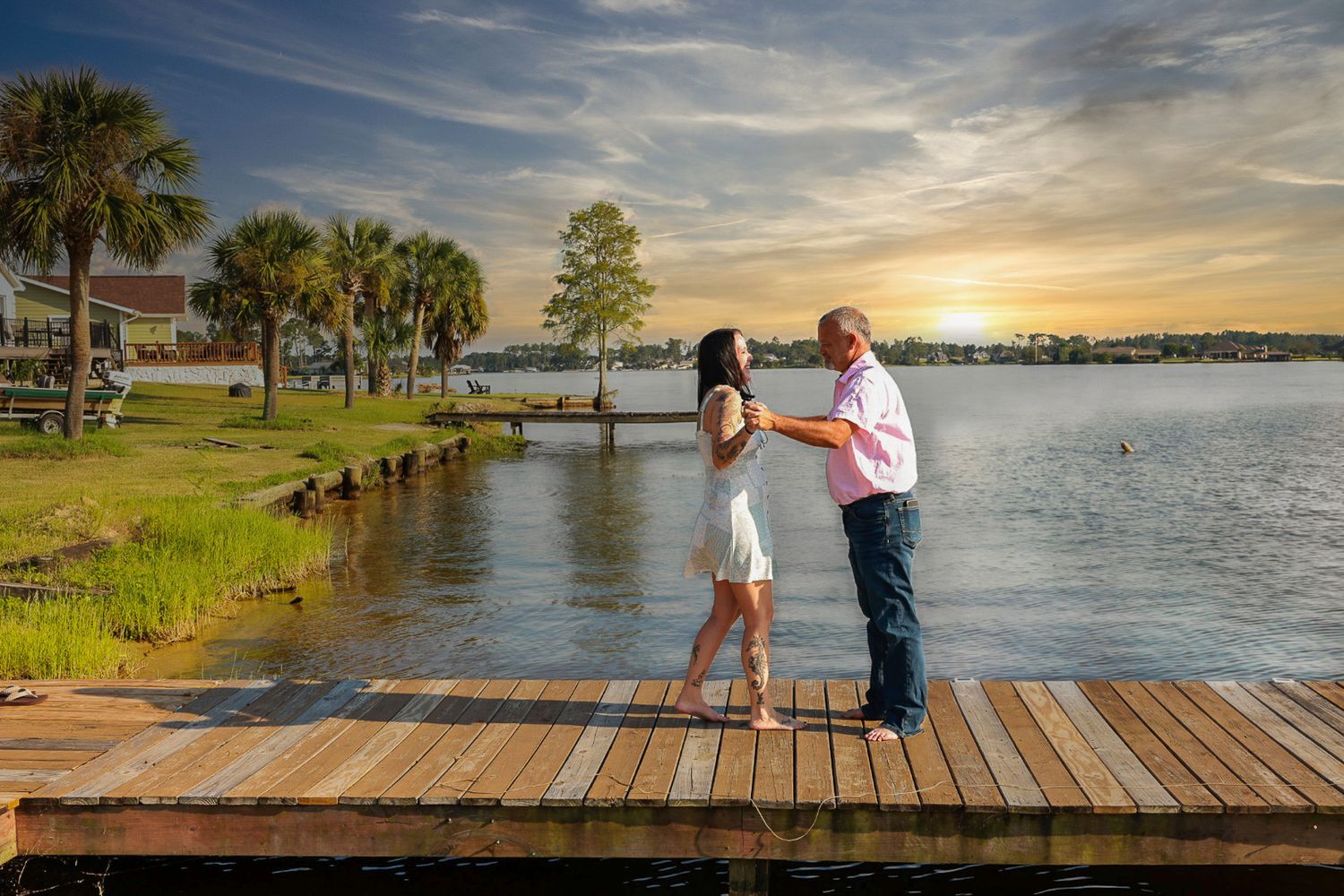 Couple holds hands and dances on a wooden lakeside dock at sunset with palm trees and calm water behind them.