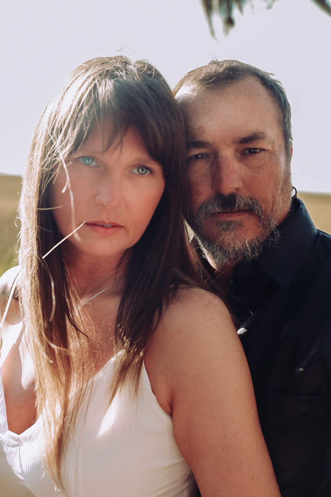 Portrait of a woman with long hair and blue eyes in a white dress beside a bearded man in a dark shirt.