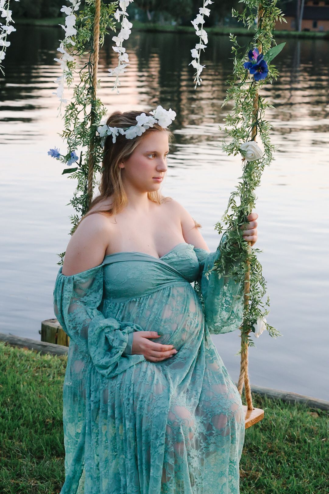 Pregnant woman in a teal lace gown and white floral crown sitting on a vine-covered rope swing beside a calm lake.