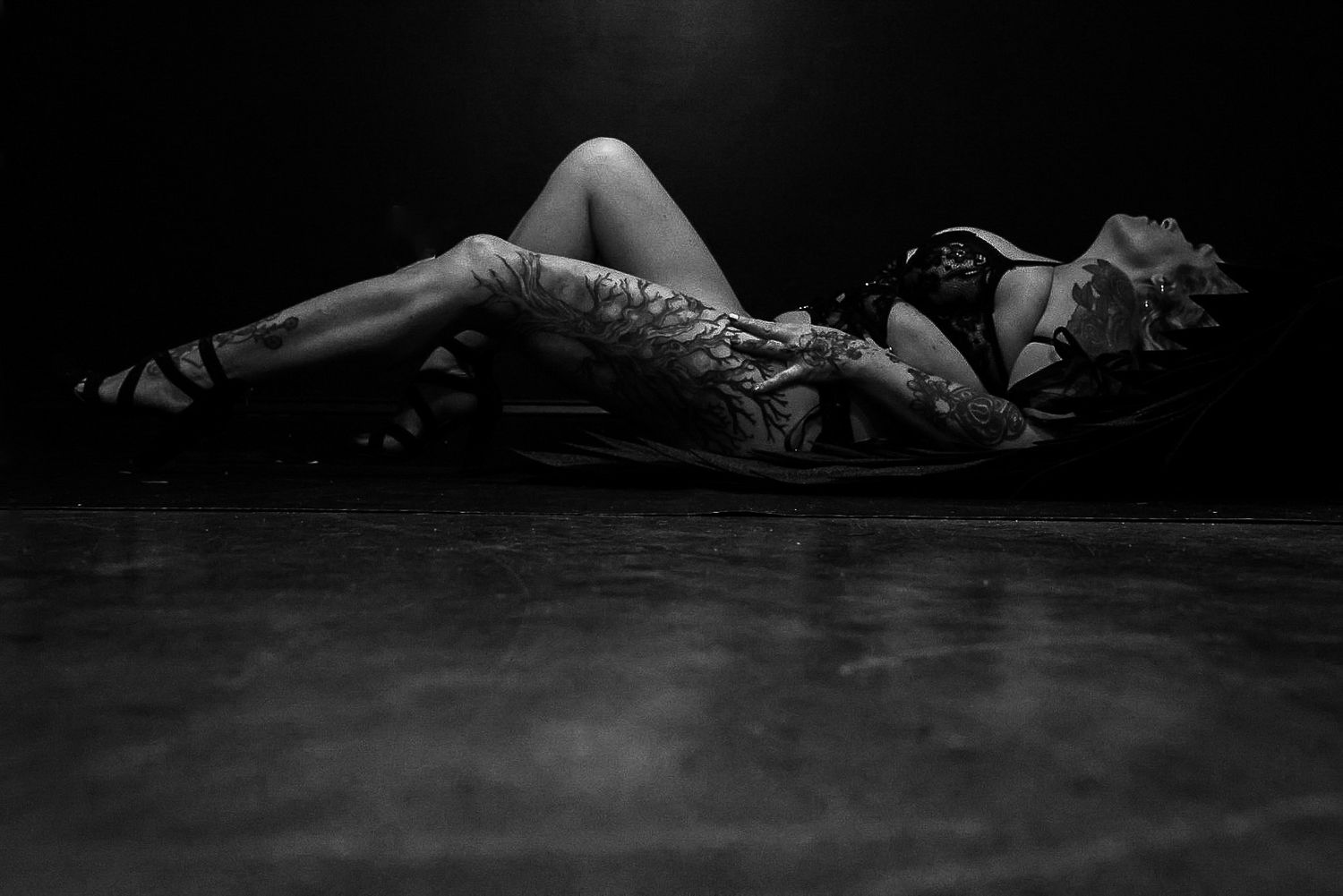 Black and white boudoir portrait of a heavily tattooed woman reclining in heels against a dark studio backdrop.