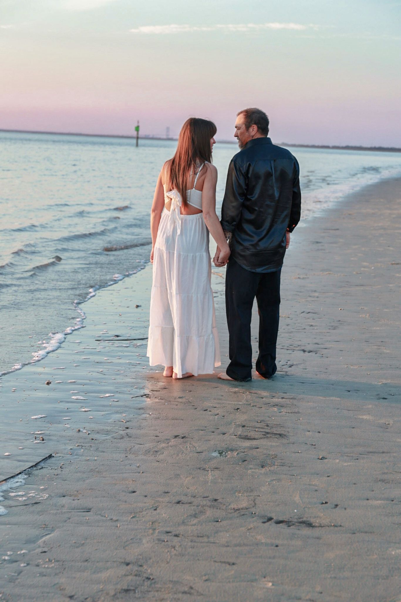 A couple holds hands while walking along a quiet shoreline at dusk, the woman in a long white dress and the man in black.