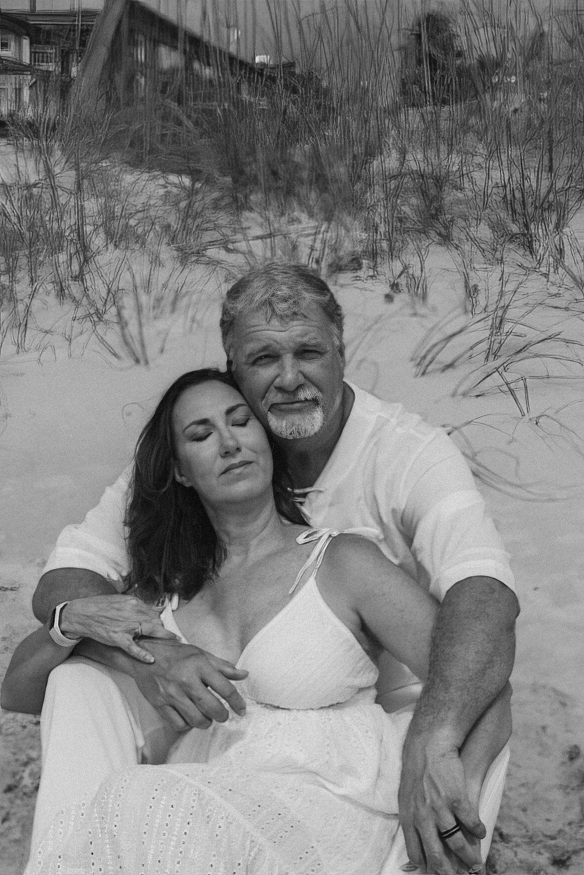 Black and white photo of a couple embracing on a sandy beach in front of dune grass, the woman resting against the man.