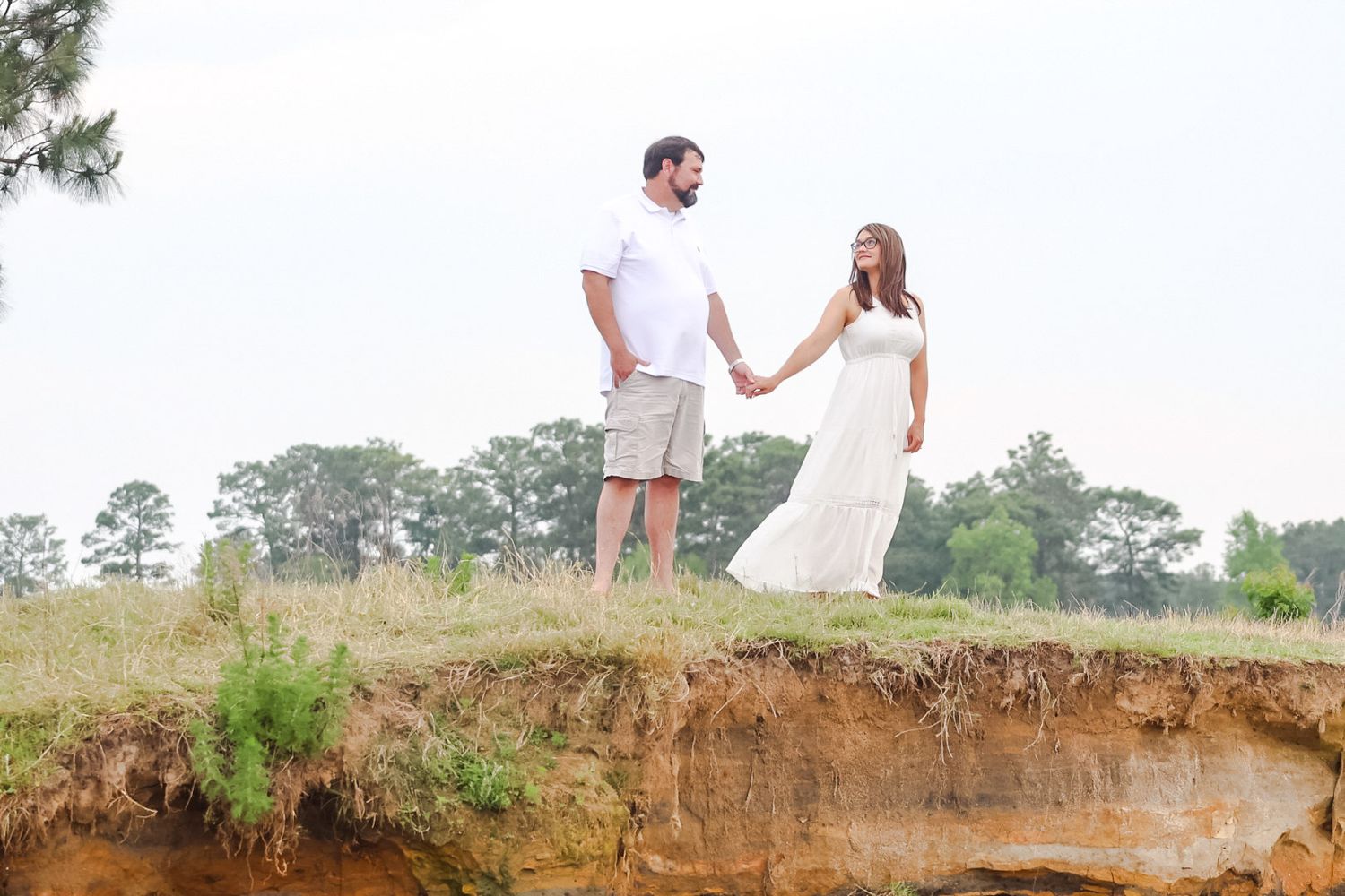 A couple holds hands while standing on a clay bluff edge, the woman in a long white dress, with trees and hazy sky behind.