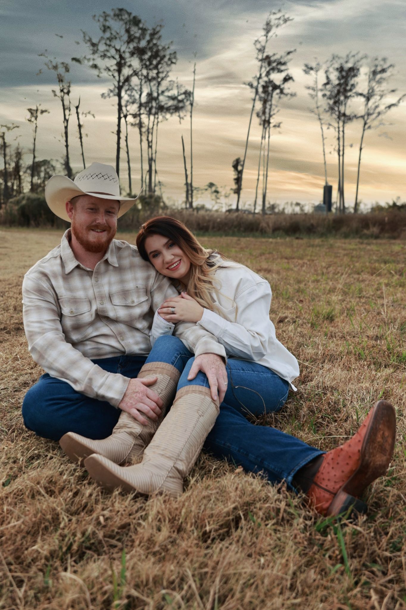 A smiling couple in cowboy hats and boots sit close together on golden grass during a sunset rural engagement photo session.