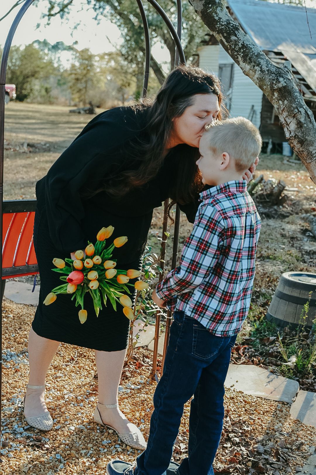 A woman in a black dress kisses a young boy in a plaid shirt on the forehead while holding a bouquet of yellow tulips.