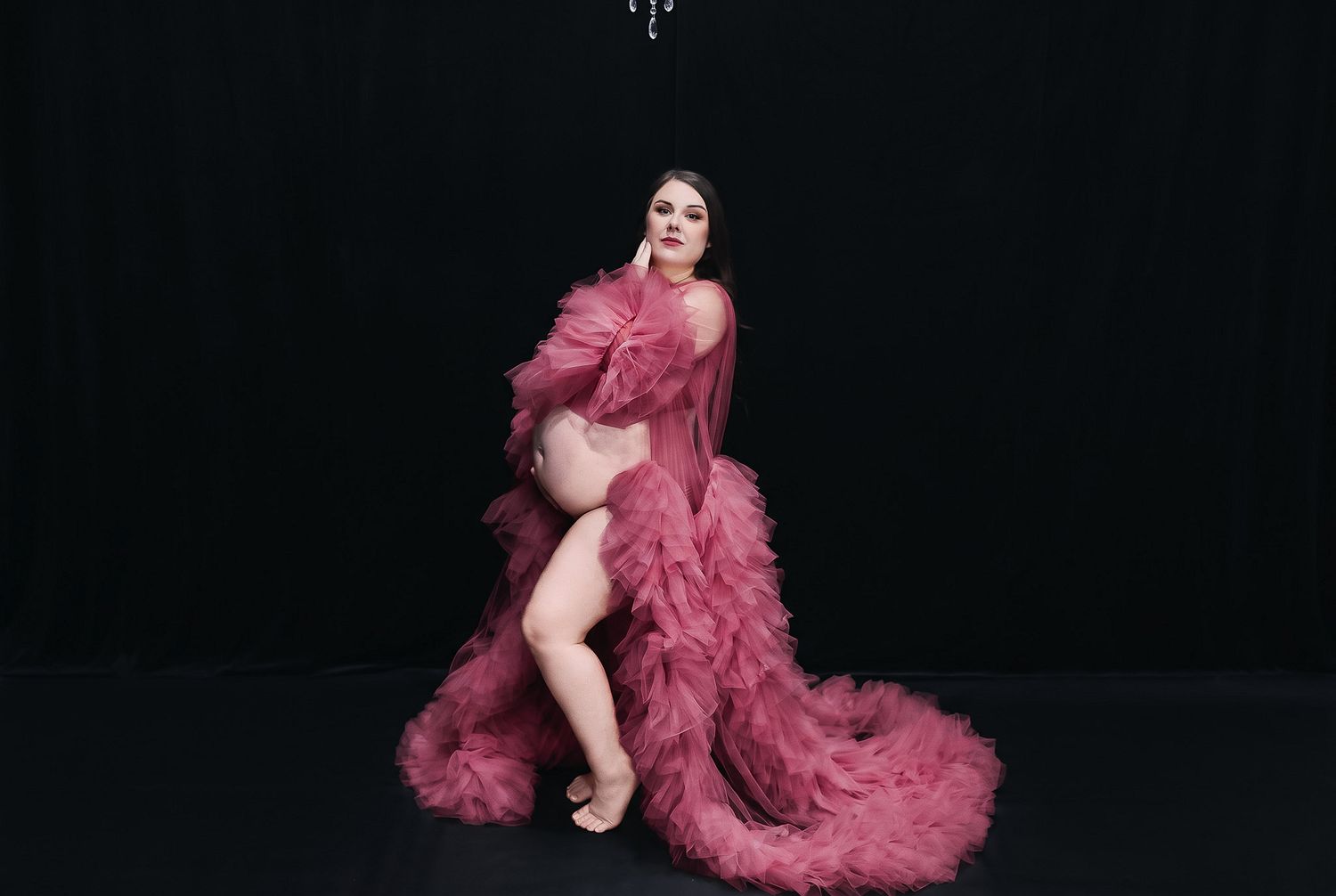 Pregnant woman poses in a dramatic pink tulle ruffled robe with a long train against a dark black backdrop in a maternity photo session.