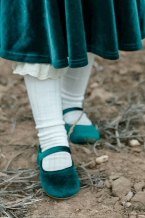 Close-up of a child's green velvet mary jane shoes, white ribbed socks and velvet skirt on dry, rocky ground.