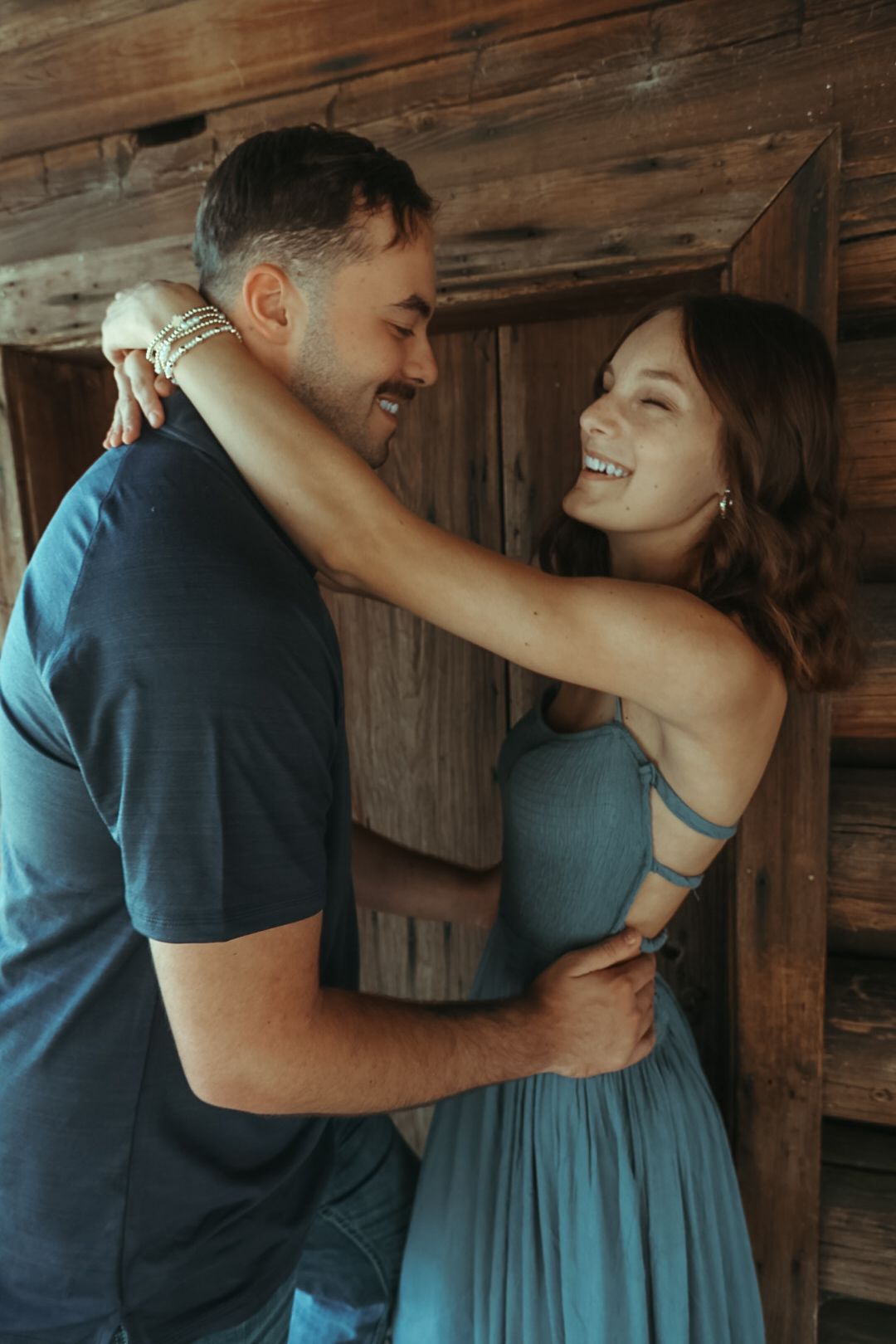Smiling couple laughing and hugging in a joyful engagement-style portrait beside a rustic wooden barn wall.