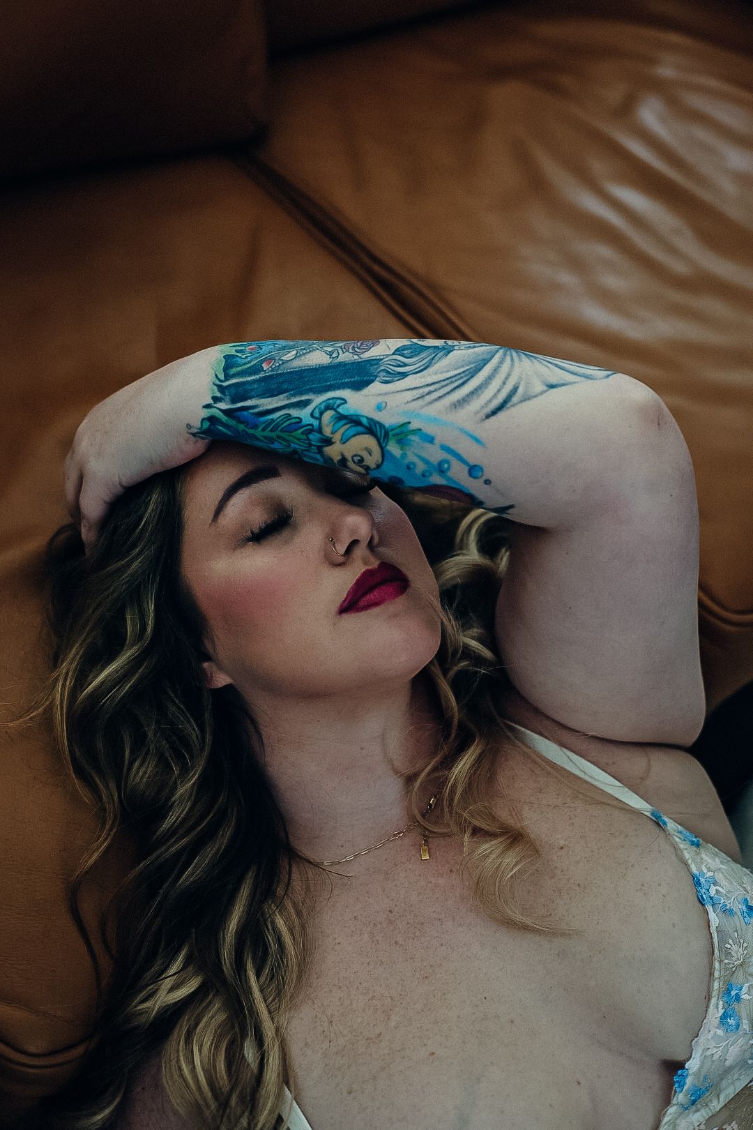 Tattooed woman with wavy hair and red lipstick rests her arm over her head on a brown leather couch.
