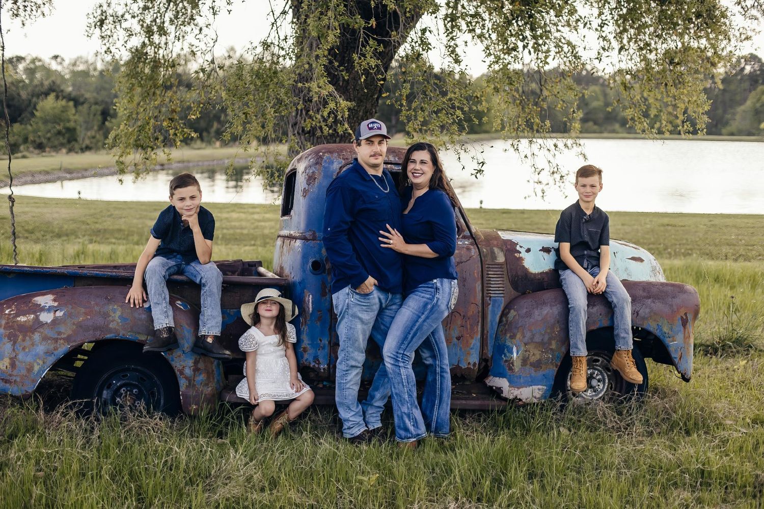 Family of five poses on and around a rustic vintage blue pickup truck in a grassy field beside a peaceful lake.