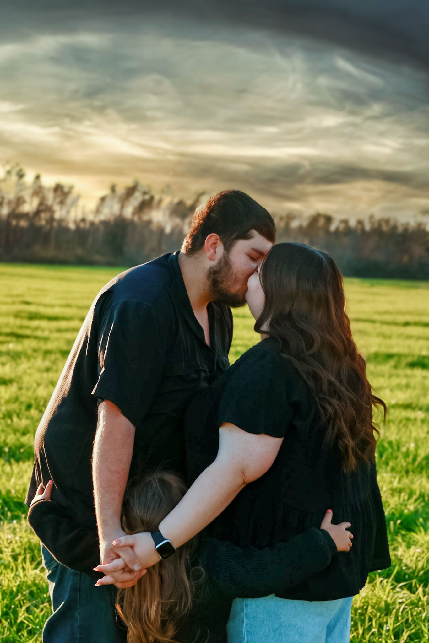 Couple kisses while embracing their child in a green field beneath a dramatic sunset sky.