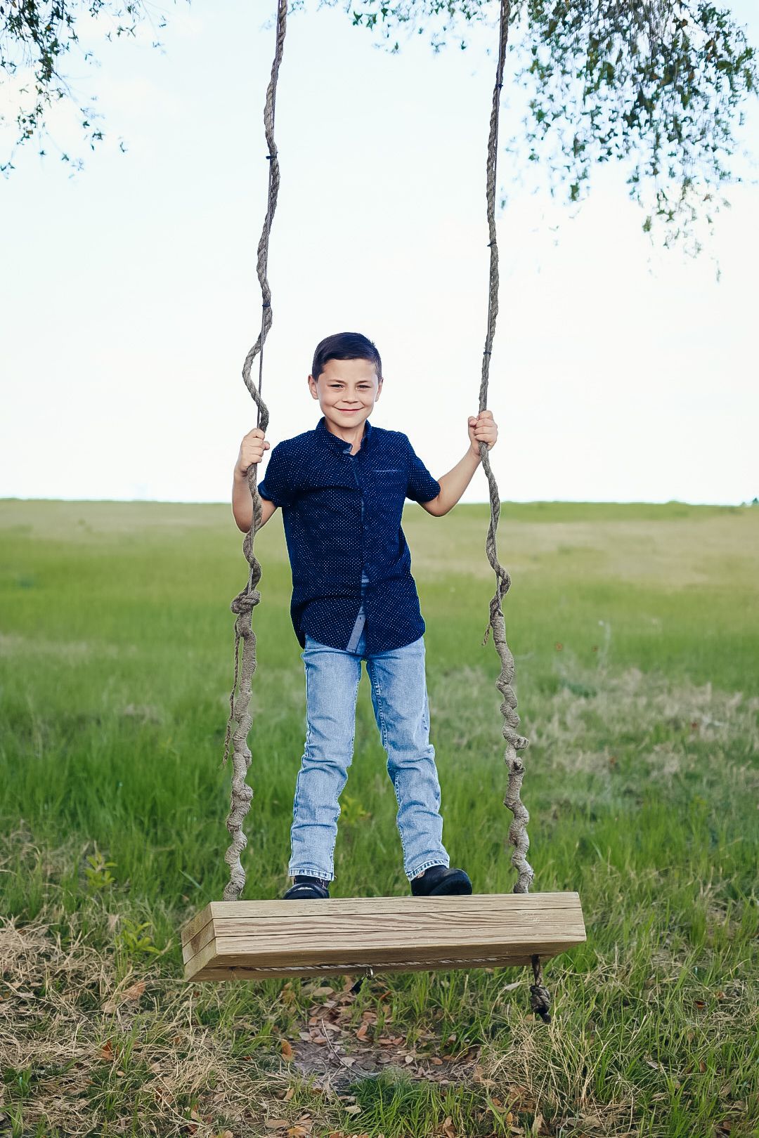 Smiling boy in a navy shirt and jeans standing on a rustic wooden rope swing in a green grassy field.