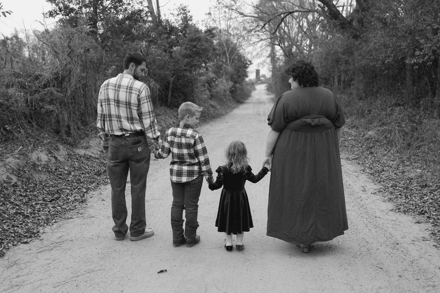 Black and white photo from behind of a family of four walking hand in hand down a country dirt road lined with trees.