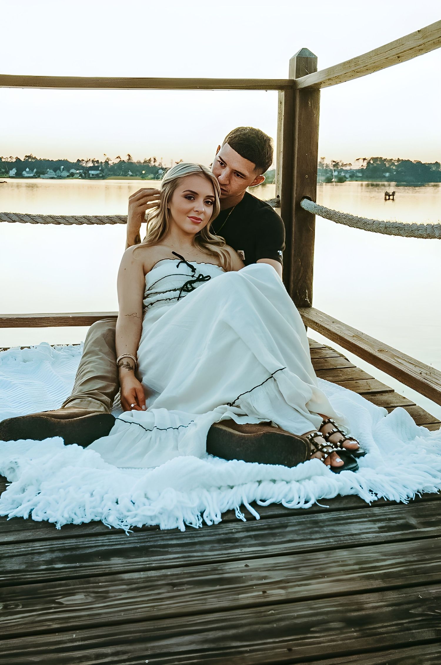 Pregnant woman in a white dress rests against her partner on a lakeside wooden dock draped with a fringed white blanket at sunset.