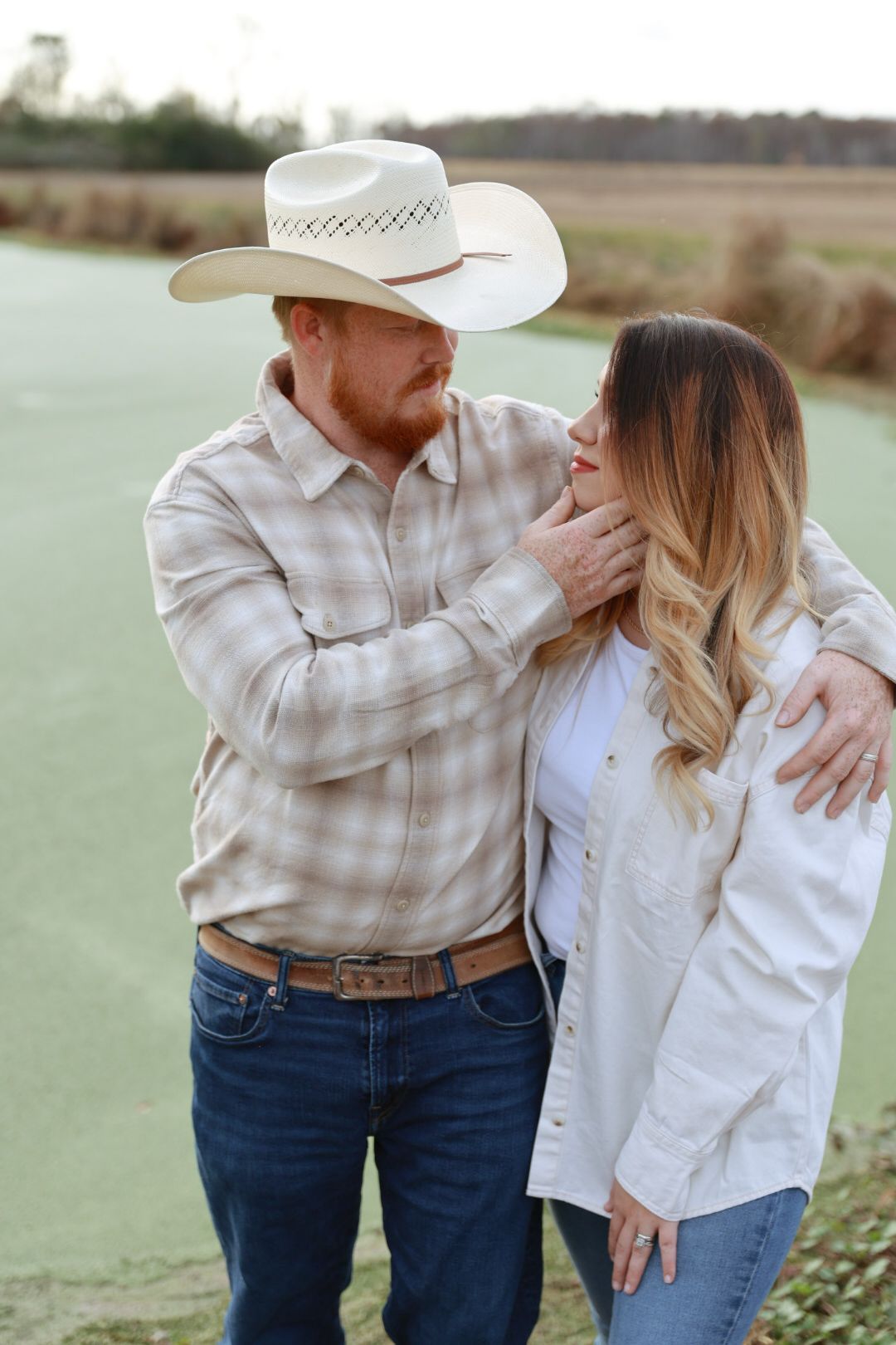 A man in a cowboy hat and plaid shirt gently touches the face of a woman in a white jacket beside a green pond.