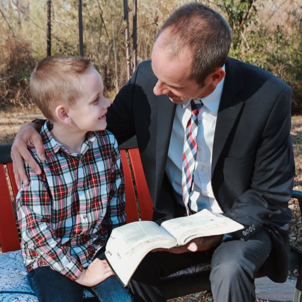 A smiling boy in a plaid shirt sits on a red bench beside a man in a suit who reads an open book outdoors.