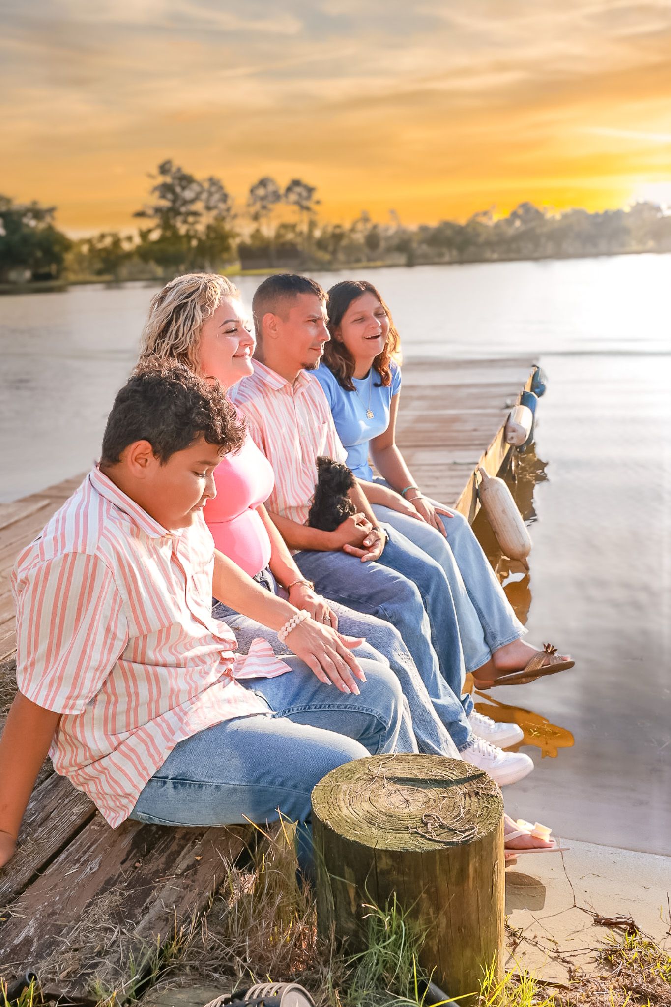 Family of four sits together on a wooden pier at golden hour with a small dog, with trees and a lake behind them.