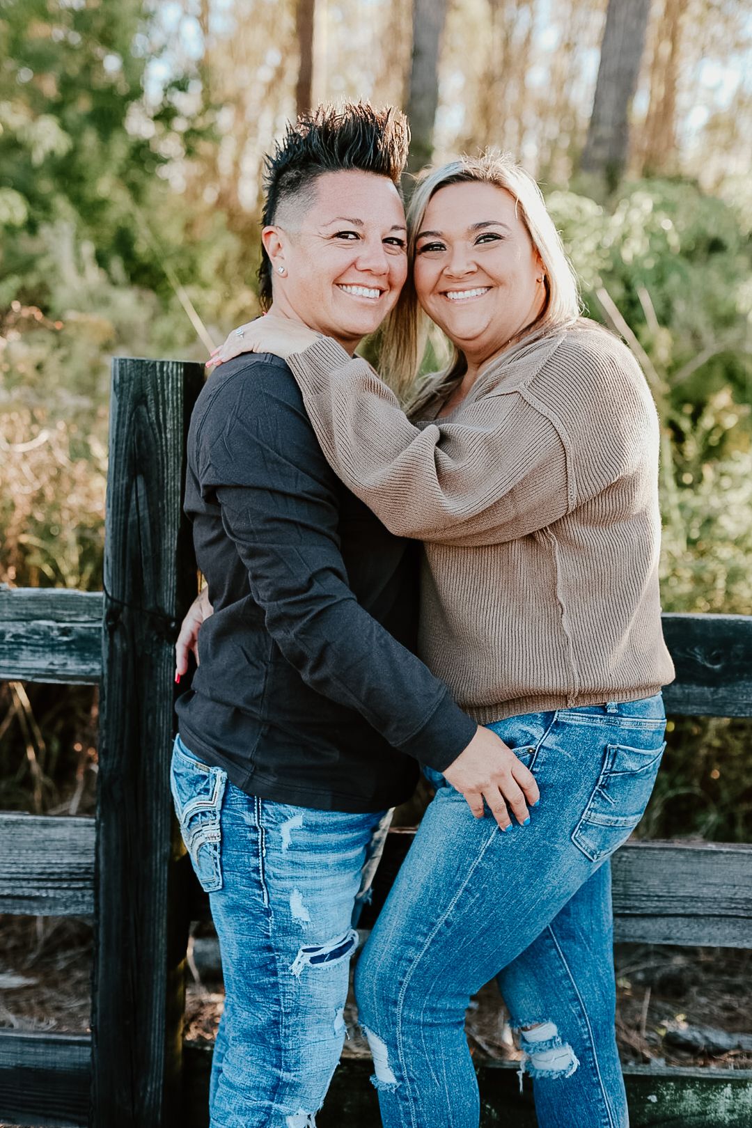 Two women in jeans and sweaters smile and embrace in front of a rustic wooden fence with trees in the background.