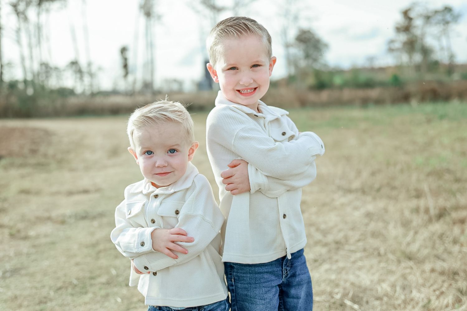Two young boys in matching cream shirts stand back to back with arms crossed, smiling in a grassy field.