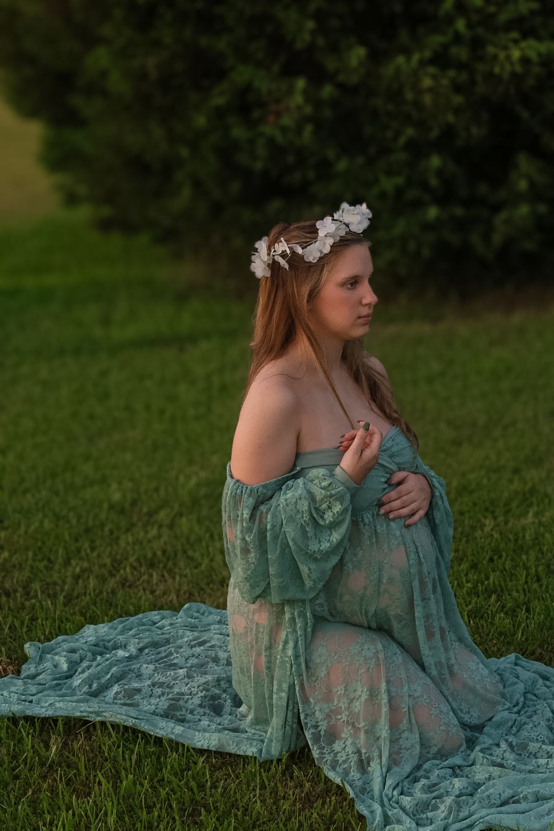 Pregnant woman wearing a white flower crown and a green lace gown kneels on grass during an outdoor maternity photo session.