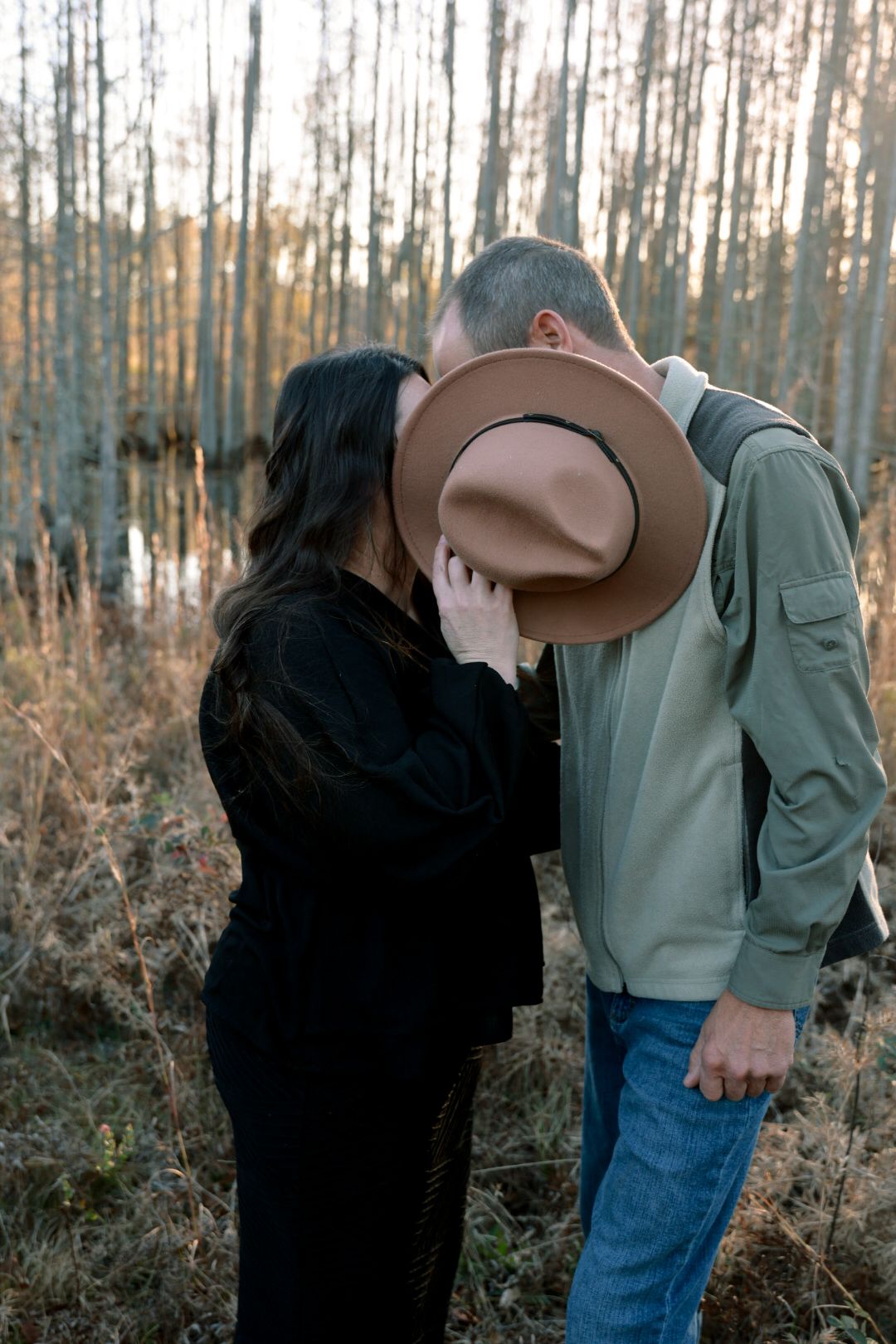 Couple shares a hidden kiss behind a brown felt hat while standing in tall golden grass near bare winter trees.