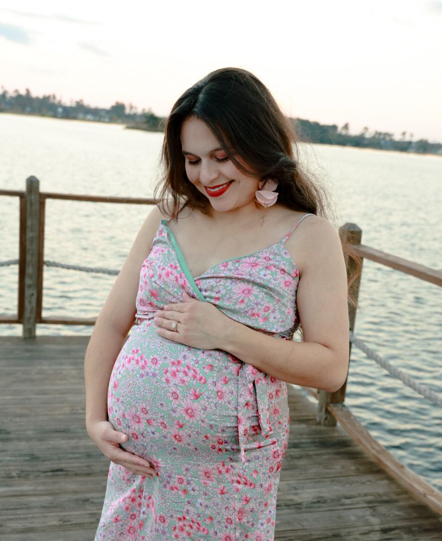 Smiling pregnant woman in red lipstick and a pink floral dress cradles her baby bump on a wooden dock overlooking a lake.