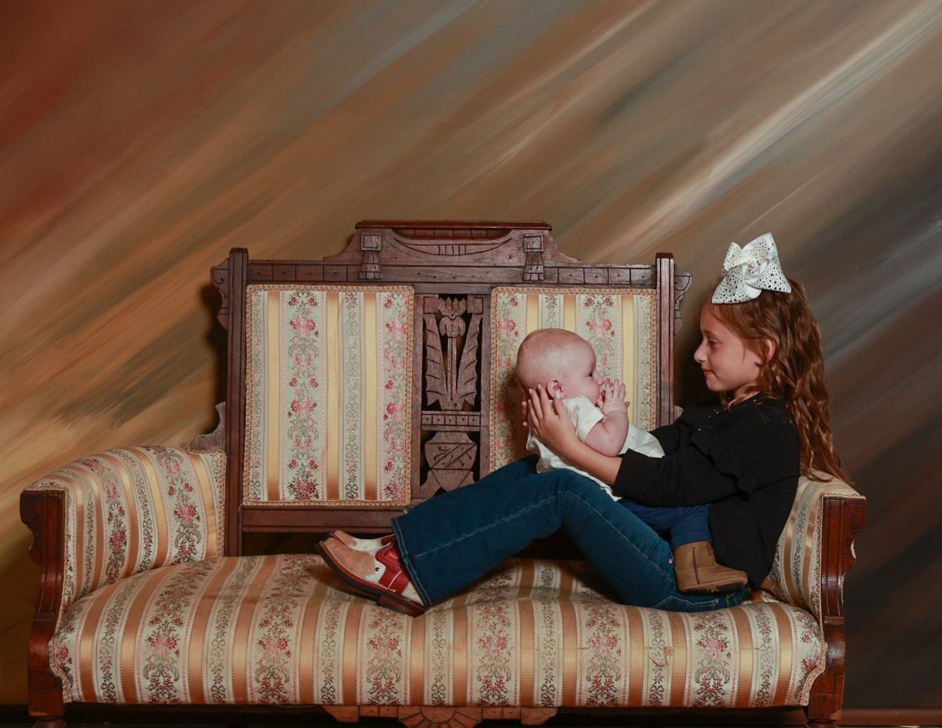Young girl with a white hair bow sits on an antique settee holding a baby in a studio family portrait.