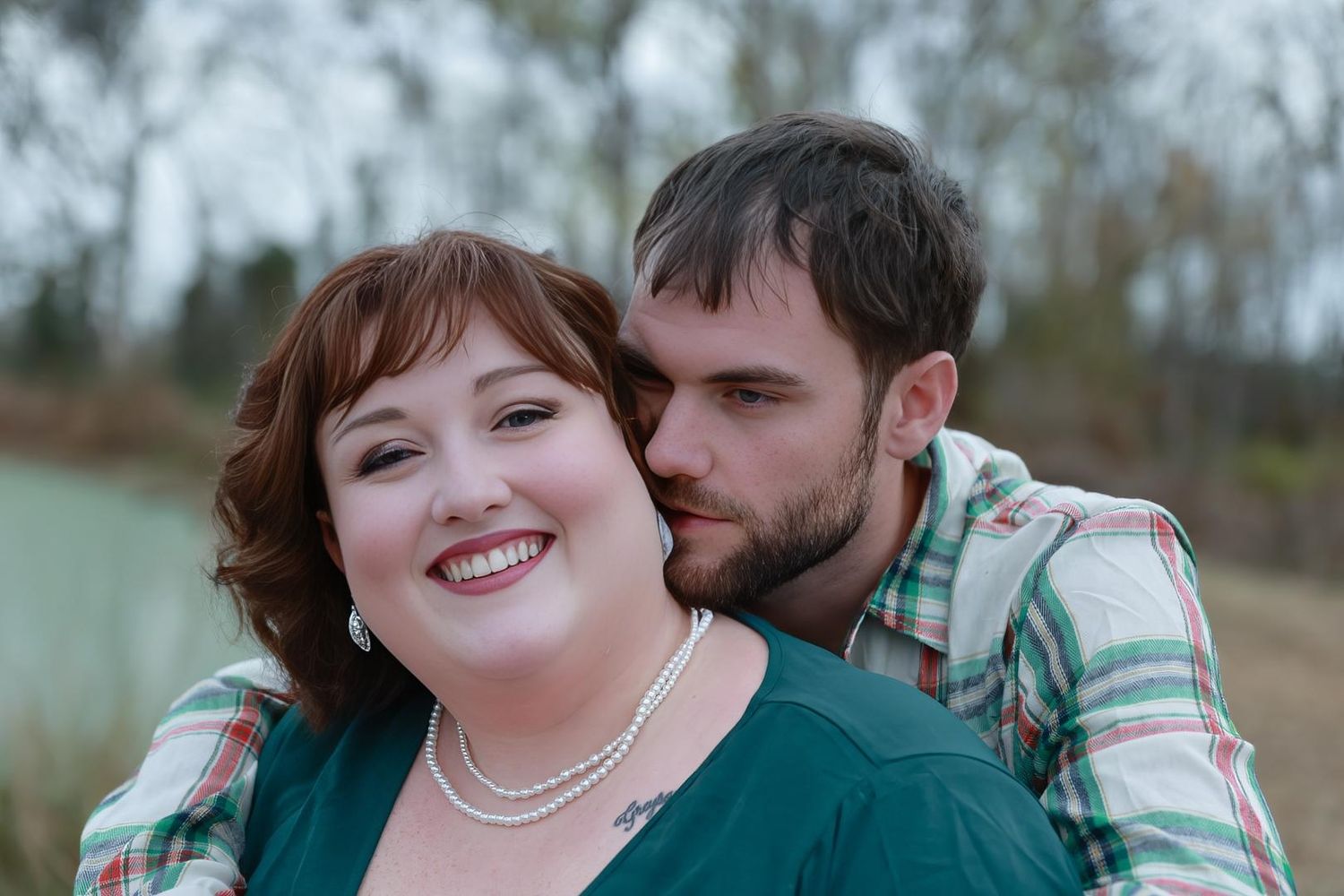 A smiling woman in a teal top and pearl necklace is hugged from behind by a bearded man in a plaid shirt by a lake.